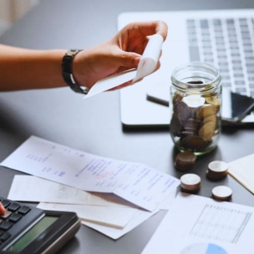 A business owner calculating expenses, surrounded by receipts and coins, signaling sudden cost increases.